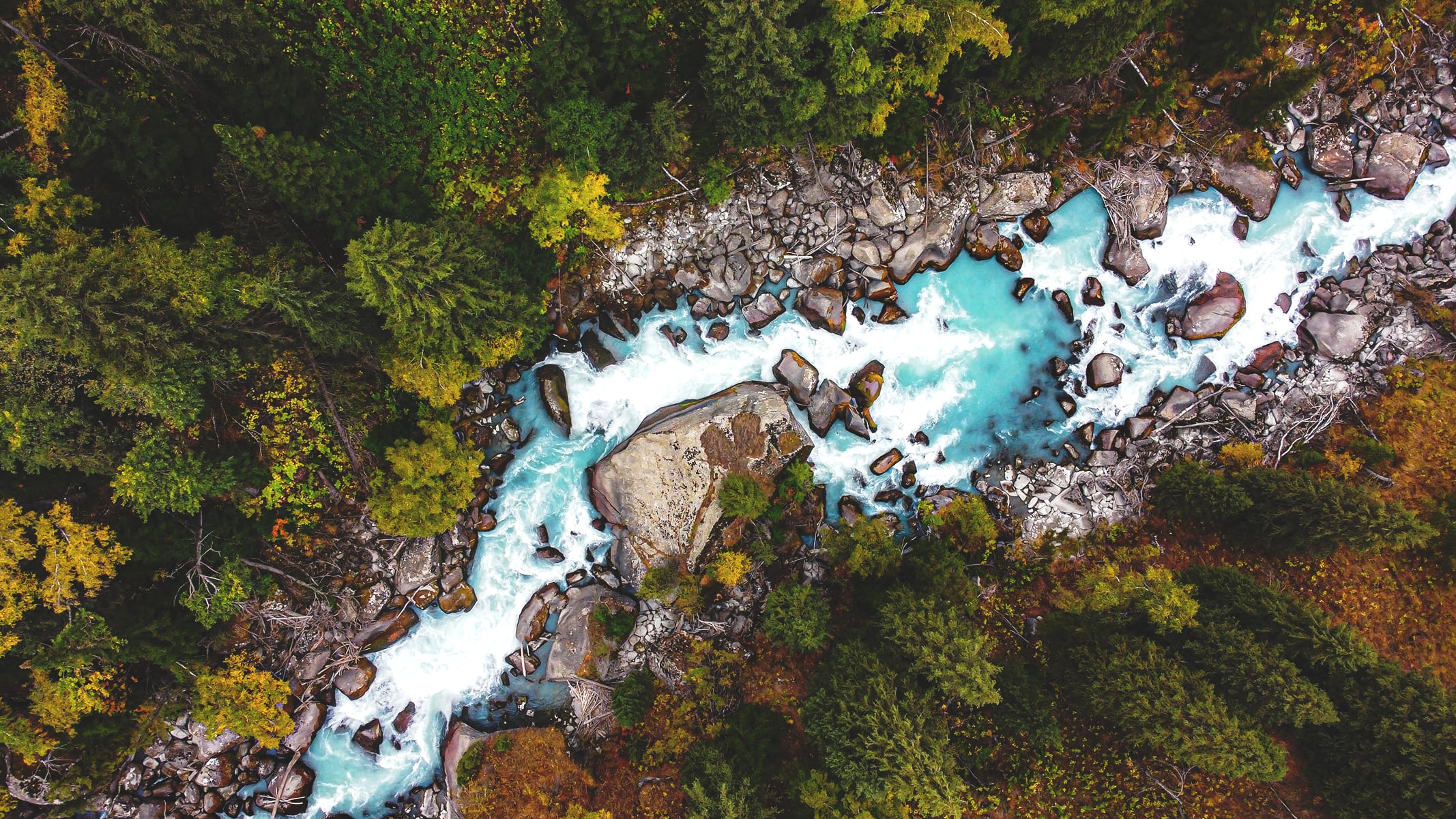 Aerial view of a river with a striking turquoise hue, flowing through a rocky terrain in a lush green forest
