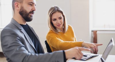 Two business professionals sit in an office setting, discussing something on a laptop which the viewer cannot see.