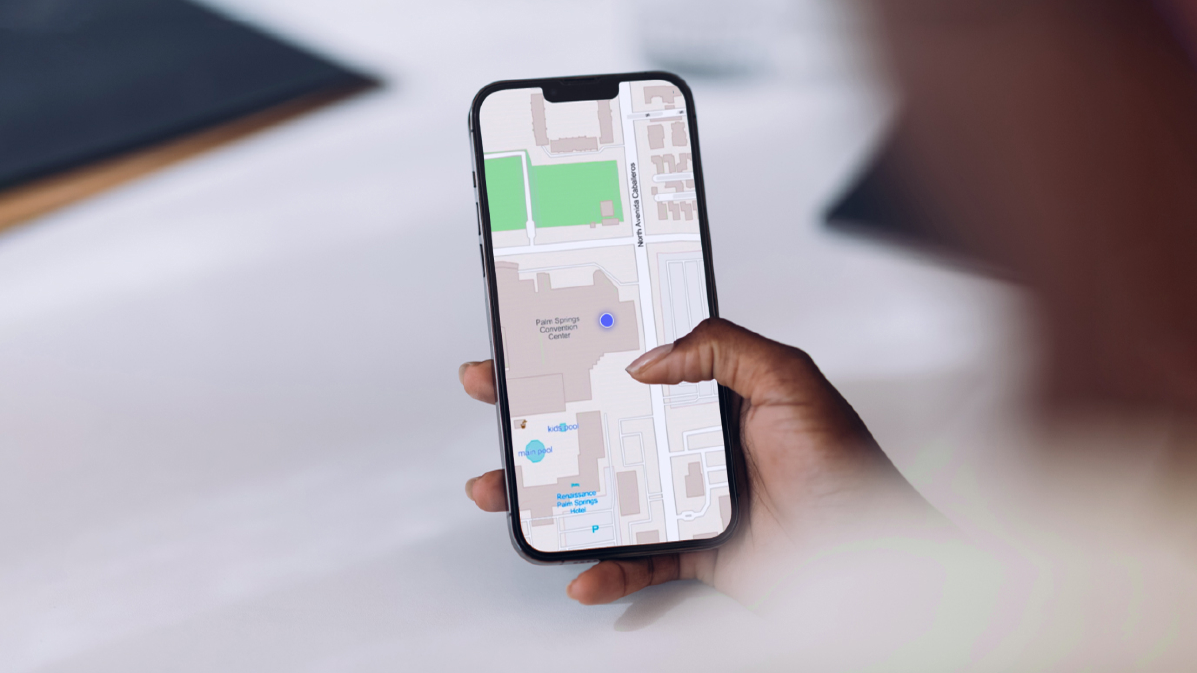 A hand holding a cell phone showing a light gray map with a blue marker on a blurred background