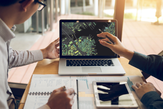 A close-up of two business professionals working at a table while looking at a laptop displaying a dark city grid map