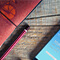 A photo of a red clothbound sketchbook, a red and white striped pencil, and a mobile phone displaying a weather forecast, all resting on a rough-grain wood tabletop