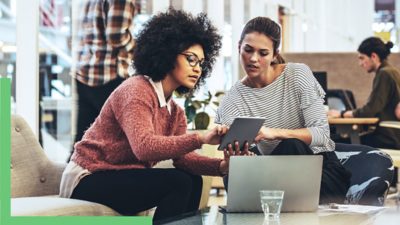 Two people seated at a low table, looking at a tablet one of them is holding and pointing to something on its screen