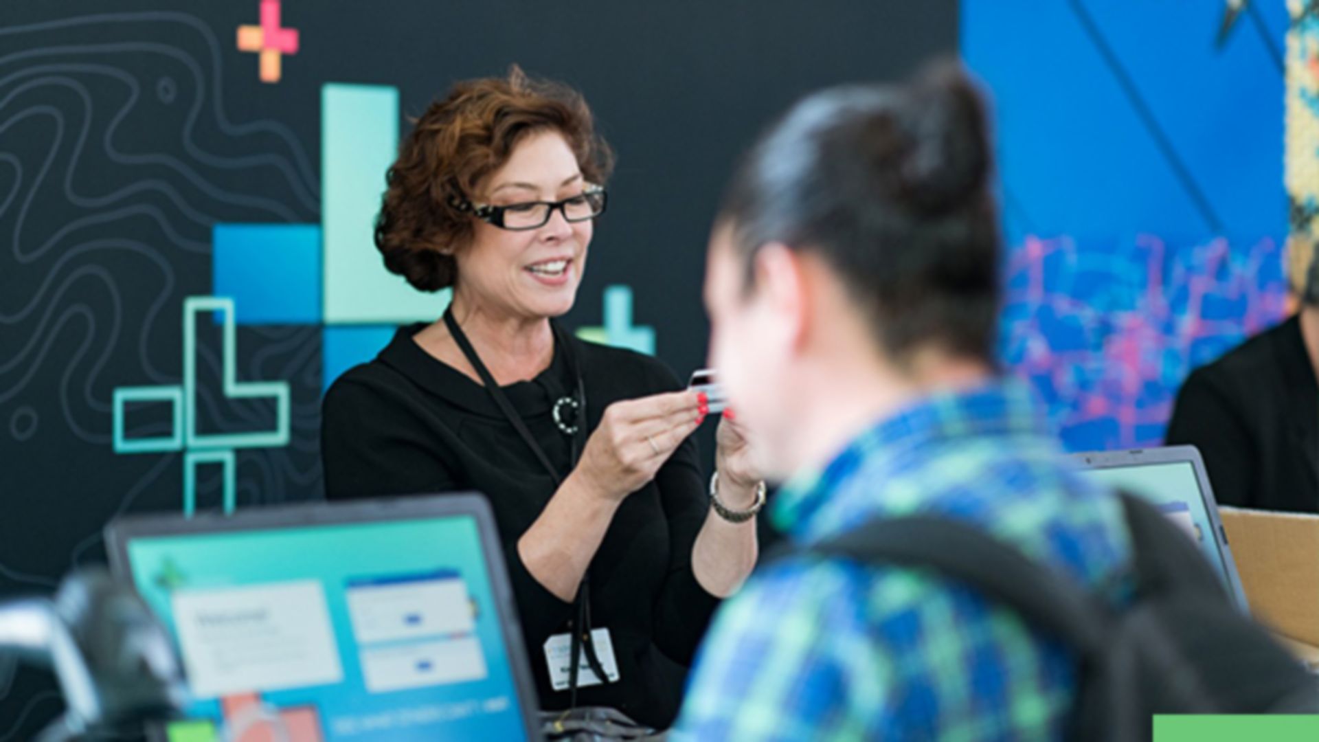 A photo of a conference staffer helping a registrant, while a person in a backpack stands in the foreground using a laptop
