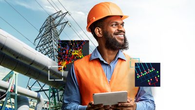 A smiling person wearing an orange safety vest and hard hat holding a tablet with an electric tower in the background