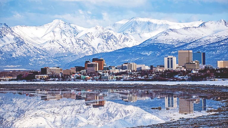 A sprawling city nestled at the base of soaring blue snow-laden mountains under a cloudswept sky
