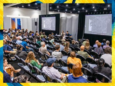 Audience seated in rows watching a presentation with large screens showing detailed maps and GIS content.