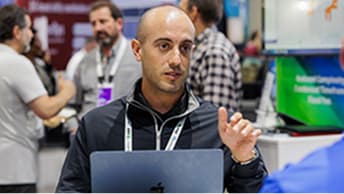 Person wearing a conference badge gestures while seated at an Expo booth with an open laptop