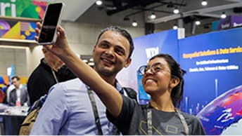 Two attendees take a selfie with a smartphone in front of a booth featuring a vibrant map-themed display and signage