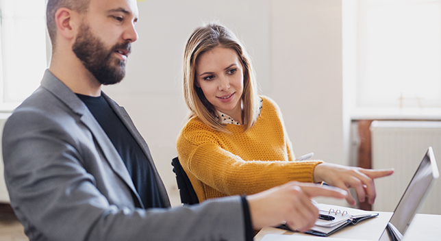 Due professionisti del business siedono in un ufficio e discutono di qualcosa su un computer portatile che lo spettatore non può vedere.