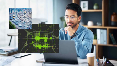 Man wearing glasses and a blue shirt sitting at his desk looking at two computer screens showing a map of a city with green dots and a photo of a busy highway