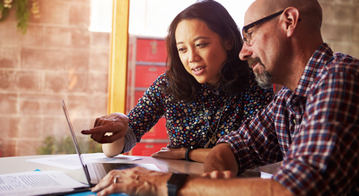 A photo of two casually-dressed people sitting in a sunlit office at a table discussing a laptop display with a brick wall in the background