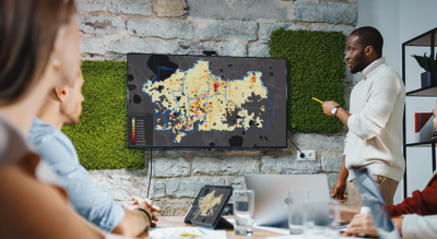 A photo of a casually dressed person in a modern conference room standing beside a wall monitor gesturing to the displayed map for a group of four seated observers in the foreground